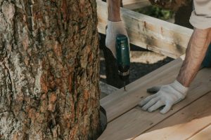 Worker drilling wooden planks on deck beside tree using power tool. DIY construction project.